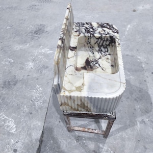 May include: A marble sink with a rectangular basin and a back splash. The sink has a white and brown marbled pattern. It is supported by a small, weathered, wooden frame. The sink's edges are fluted.