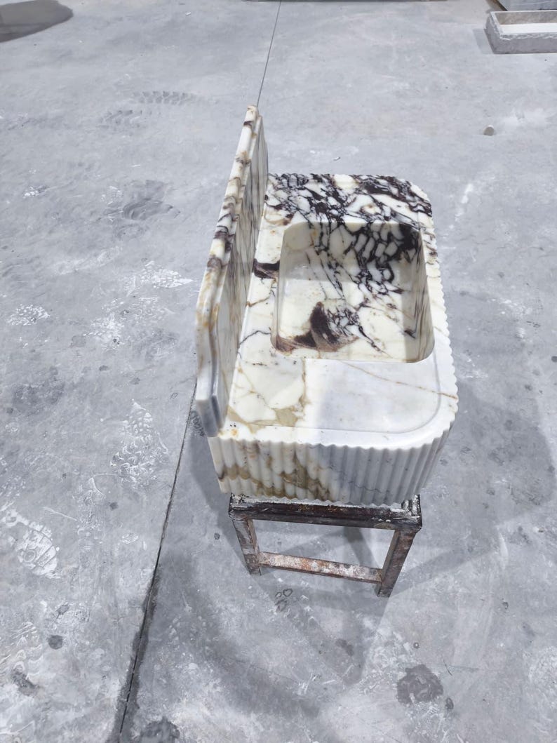 May include: A marble sink with a rectangular basin and a back splash. The sink has a white and brown marbled pattern. It is supported by a small, weathered, wooden frame. The sink's edges are fluted.