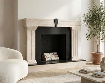 May include: A beige stone fireplace with a black interior and a black accent piece. The fireplace has a stack of birch logs inside. A potted olive tree and a white chair are in the background. A coffee table with books is in front of the fireplace.