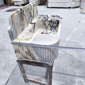 May include: A marble sink with a rectangular basin and a decorative back. The sink features a white and brown veined pattern. A measuring tape shows the sink is approximately 22 inches long. The sink is placed on a wooden stand.