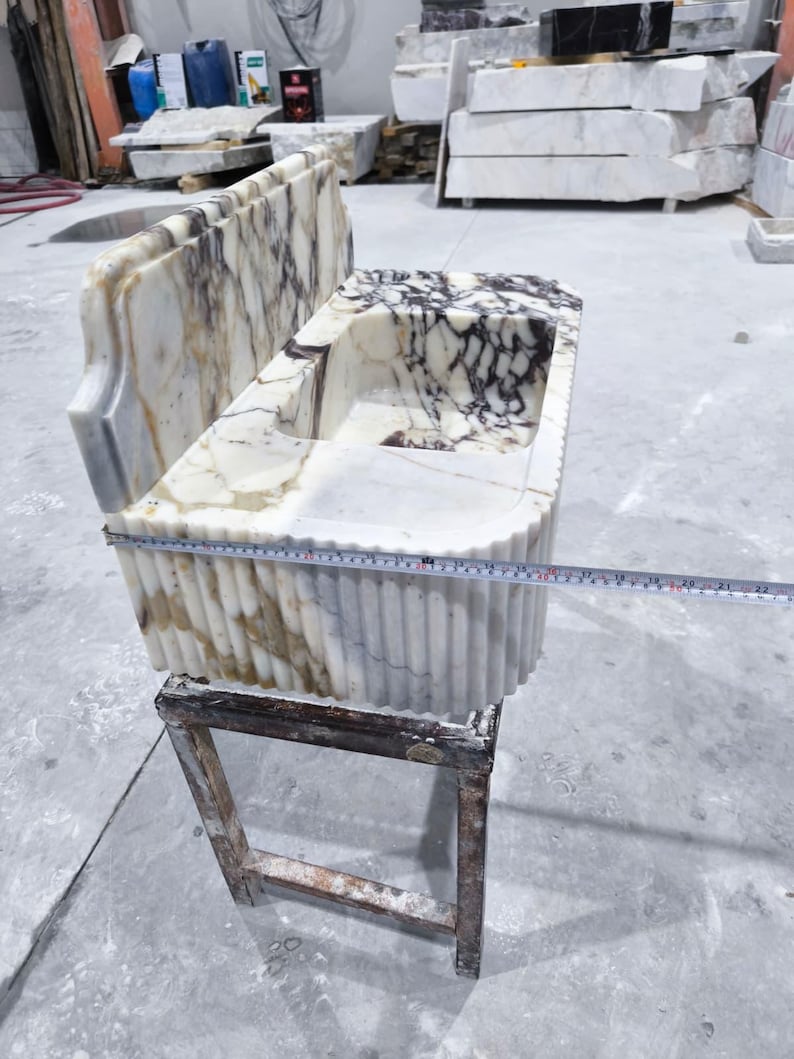 May include: A marble sink with a rectangular basin and a decorative back. The sink features a white and brown veined pattern. A measuring tape shows the sink is approximately 22 inches long. The sink is placed on a wooden stand.