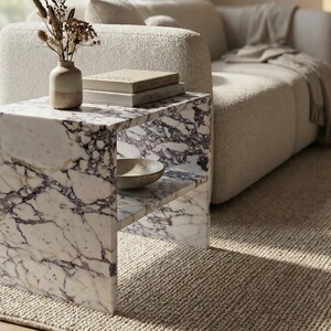 May include: A marble side table with a white and purple veined pattern. The table has a shelf and is next to a beige sofa. A vase with dried plants and books are on top of the table. A small bowl is on the shelf.