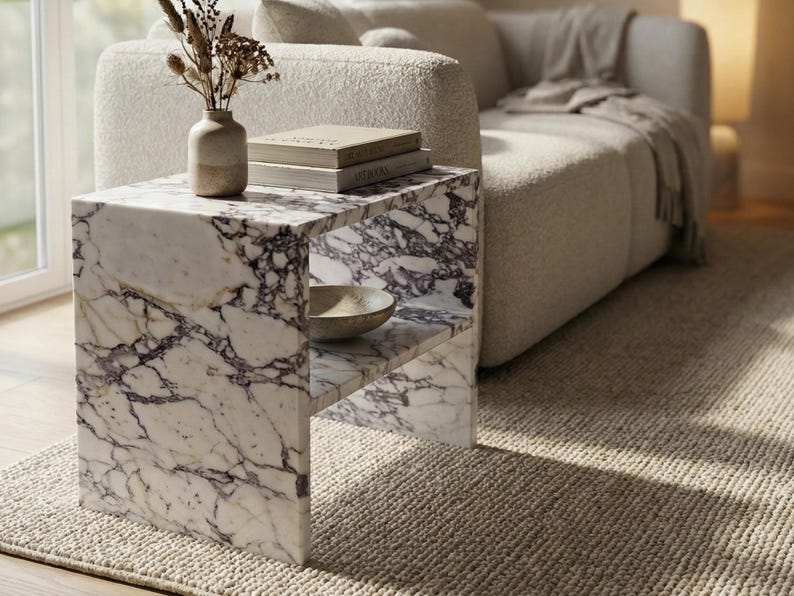May include: A marble side table with a white and purple veined pattern. The table has a shelf and is next to a beige sofa. A vase with dried plants and books are on top of the table. A small bowl is on the shelf.