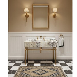 Puede incluir: Interior de ba&ntilde;o con lavabo de m&aacute;rmol sobre soporte dorado, grifer&iacute;a dorada y espejo. Las paredes beige tienen apliques. Una alfombra estampada y un suelo ajedrezado en blanco y negro completan el dise&ntilde;o.