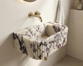 May include: A wall-mounted rectangular bathroom sink crafted from white marble with prominent dark purple veining. The exterior features a vertical fluted texture, and the basin is paired with a brushed gold wall-mounted faucet fixture.