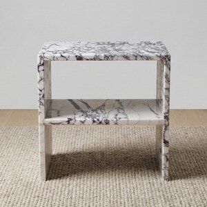 May include: A white marble side table with a shelf. The table has a rectangular shape with a dark gray veining pattern throughout. The table is made of solid marble and has a modern design.