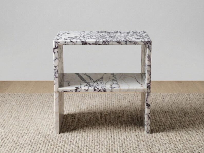 May include: A white marble side table with a shelf. The table has a rectangular shape with a dark gray veining pattern throughout. The table is made of solid marble and has a modern design.