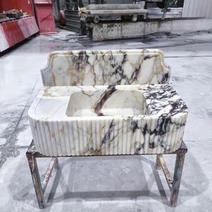 May include: A marble sink with a rectangular basin and a decorative back. The sink has a white and brown veined pattern and is supported by a metal stand. The sink's front has a fluted design.