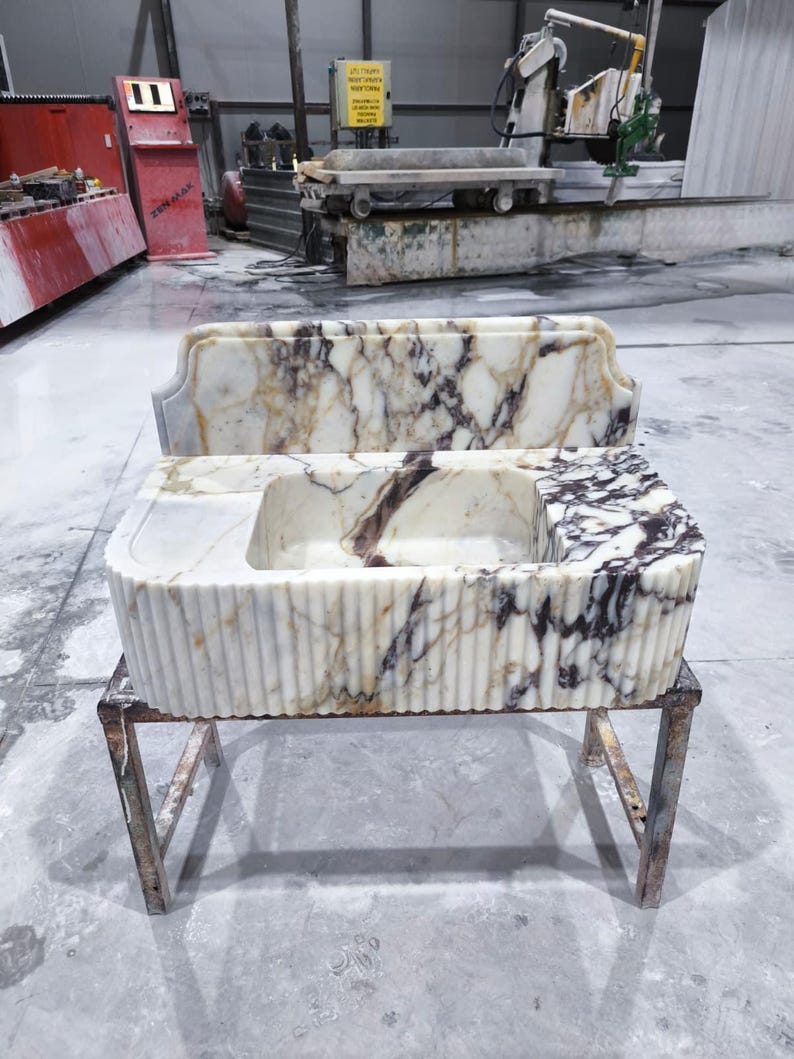 May include: A marble sink with a rectangular basin and a decorative back. The sink has a white and brown veined pattern and is supported by a metal stand. The sink's front has a fluted design.