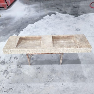 Peut inclure: Un évier rectangulaire en pierre de couleur claire avec deux bassins carrés. L'évier est soutenu par un cadre métallique avec des pieds inclinés. La pierre a une surface rugueuse et texturée. Le fond est un sol en béton.