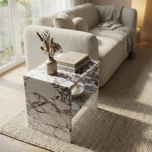 May include: A modern living room scene featuring a white sofa, a marble side table, and a beige rug. The side table has a shelf and is adorned with a vase of dried flowers, books, and a small bowl. The room is bathed in natural light.