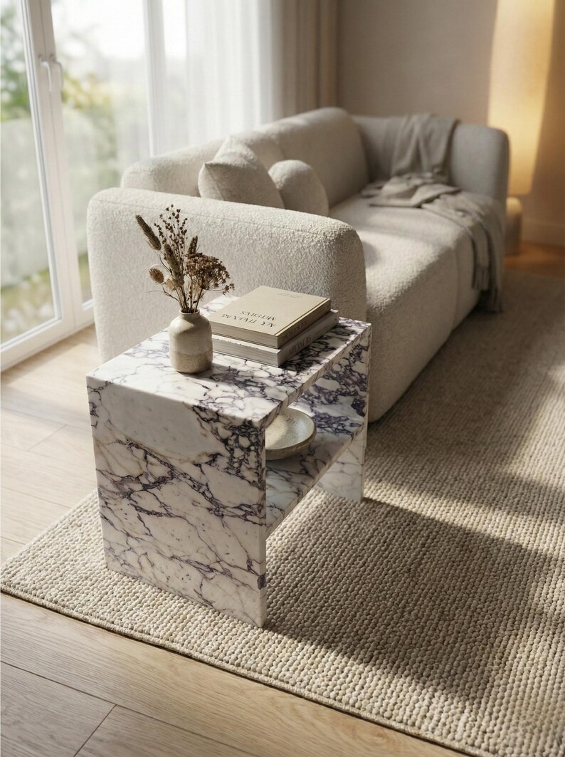 May include: A modern living room scene featuring a white sofa, a marble side table, and a beige rug. The side table has a shelf and is adorned with a vase of dried flowers, books, and a small bowl. The room is bathed in natural light.