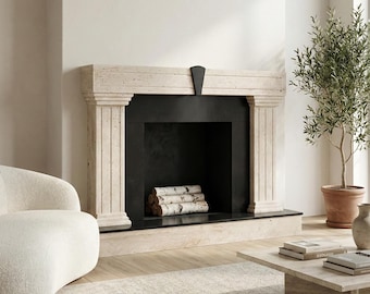 May include: A beige stone fireplace with a black interior and a black accent piece. The fireplace has a stack of birch logs inside. A potted olive tree and a white chair are in the background. A coffee table with books is in front of the fireplace.