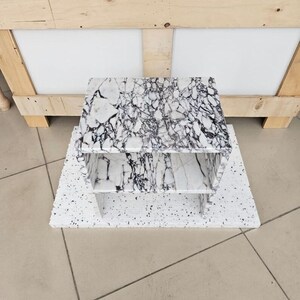 May include: A white and black marble-patterned shelf unit with two shelves. The unit sits on a speckled white base. The background includes a wooden crate and a tiled floor.