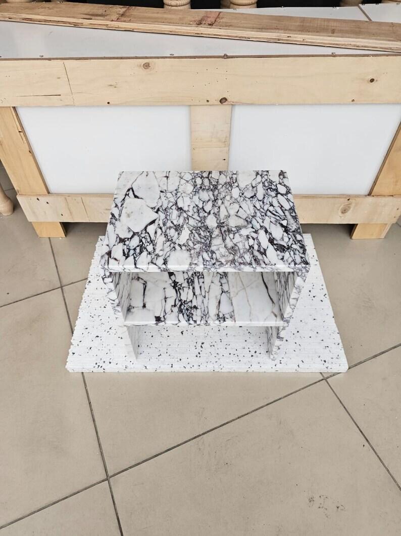 May include: A white and black marble-patterned shelf unit with two shelves. The unit sits on a speckled white base. The background includes a wooden crate and a tiled floor.
