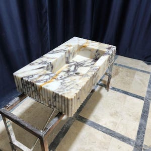 Puede incluir: Un lavabo rectangular de m&aacute;rmol con una cubeta tallada y un exterior estriado. El lavabo presenta una mezcla de vetas blancas, marrones y grises. Est&aacute; soportado por un soporte met&aacute;lico. El lavabo es un accesorio de ba&ntilde;o.