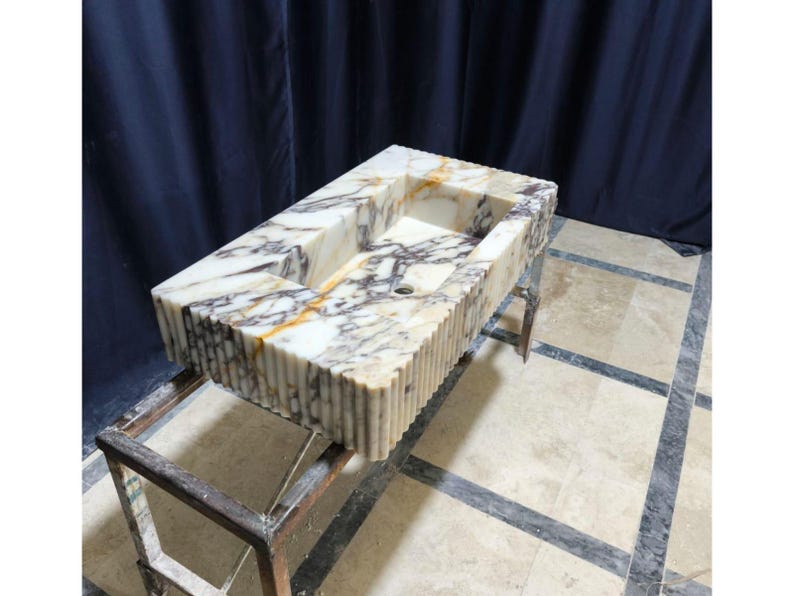 Puede incluir: Un lavabo rectangular de m&aacute;rmol con una cubeta tallada y un exterior estriado. El lavabo presenta una mezcla de vetas blancas, marrones y grises. Est&aacute; soportado por un soporte met&aacute;lico. El lavabo es un accesorio de ba&ntilde;o.