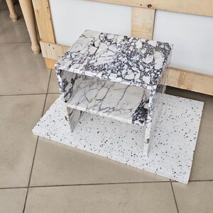 May include: A white and black marble-patterned side table with two shelves. The table sits on a white speckled base. The table is made of a marble-like material and has a modern design. The background includes a wooden structure and a tiled floor.