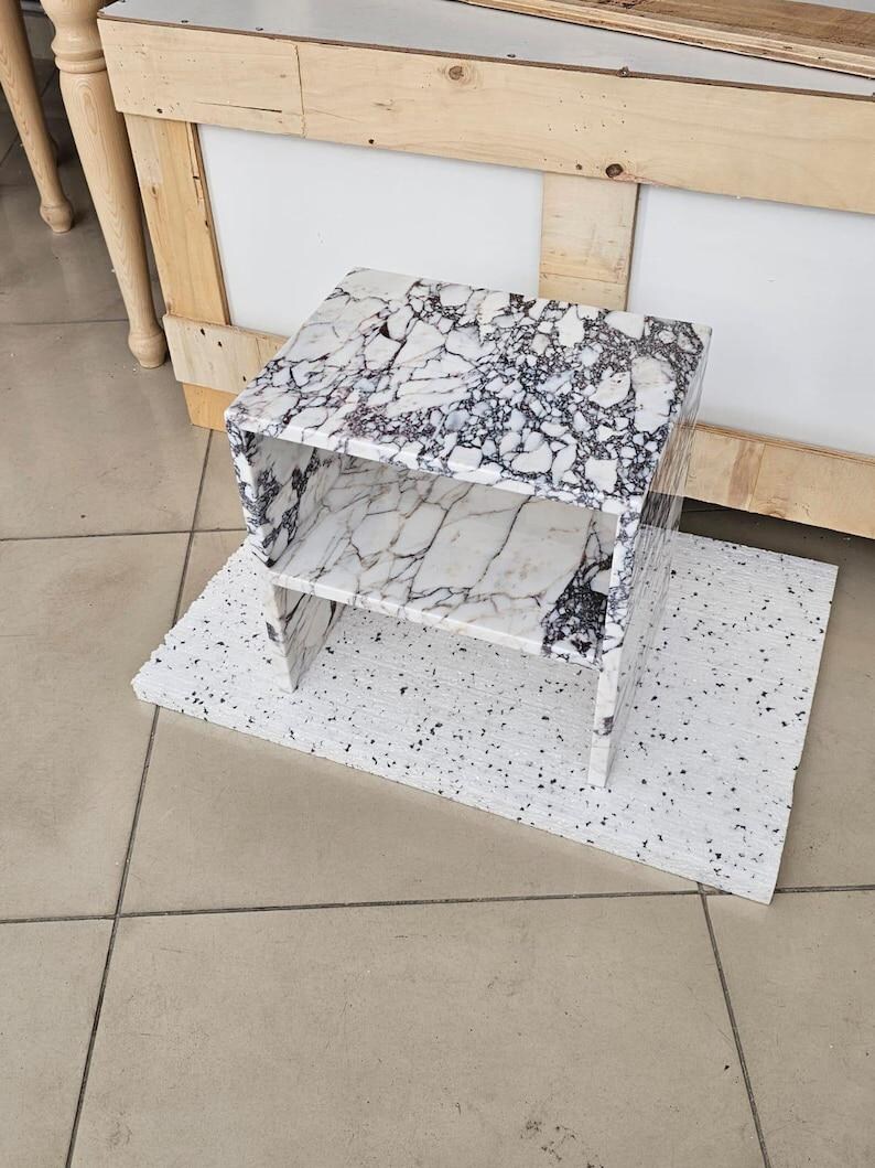 May include: A white and black marble-patterned side table with two shelves. The table sits on a white speckled base. The table is made of a marble-like material and has a modern design. The background includes a wooden structure and a tiled floor.
