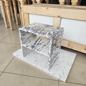 May include: A small, marble-patterned side table with a shelf, set on a white, speckled base. The table has a rectangular shape with a white and gray veined pattern. The background includes wooden furniture.