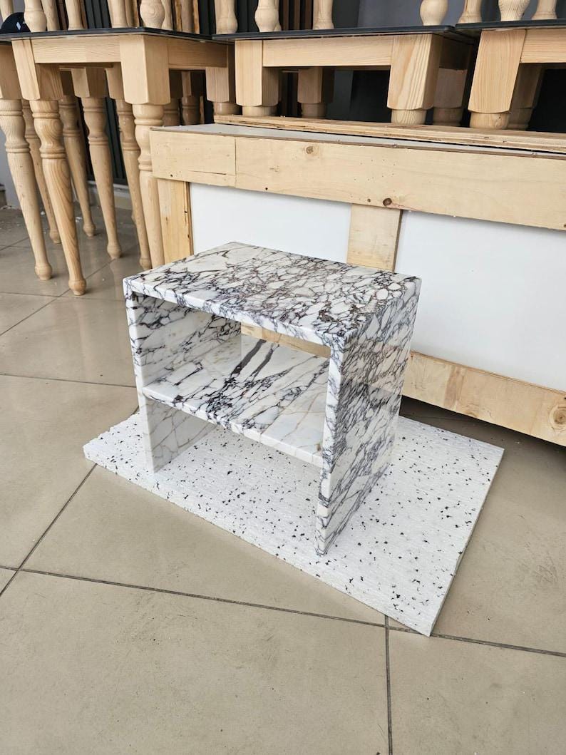 May include: A small, marble-patterned side table with a shelf, set on a white, speckled base. The table has a rectangular shape with a white and gray veined pattern. The background includes wooden furniture.