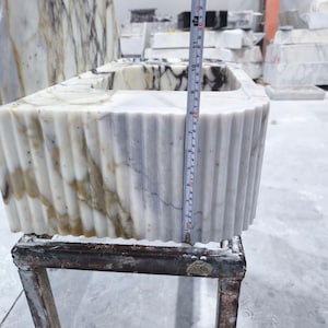 May include: A rectangular marble sink with a fluted exterior. The sink is white with brown and gray veining. The height is approximately 12 inches. The sink is sitting on a metal stand in a workshop setting.