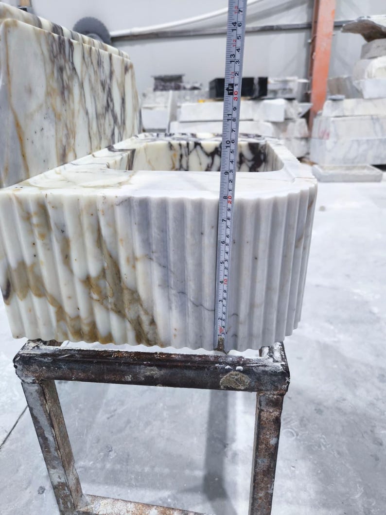 May include: A rectangular marble sink with a fluted exterior. The sink is white with brown and gray veining. The height is approximately 12 inches. The sink is sitting on a metal stand in a workshop setting.