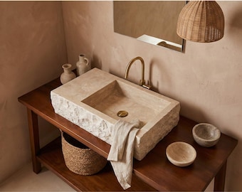May include: A stone bathroom sink with a rectangular basin and rough-hewn edges sits on a dark wood vanity. A gold faucet and drain complement the sink. A woven basket and two small bowls are on the vanity. A linen towel hangs over the sink.