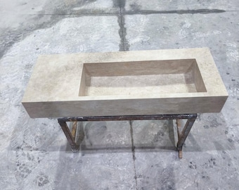 May include: A rectangular, beige stone sink with a recessed basin. The sink is supported by a metal stand with angled legs. The stone has a natural, textured appearance. The sink is designed for bathroom or kitchen use.