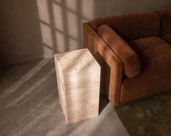 Puede incluir: Una mesa auxiliar rectangular hecha de piedra travertino beige con una superficie superior porosa y texturizada y lados lisos y veteados. La pieza presenta una paleta de colores bronceados naturales con patrones minerales visibles en toda la estructura.