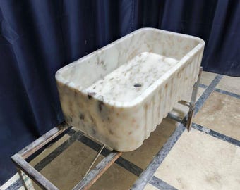 Peut inclure: Un évier rectangulaire en marbre avec un motif moucheté beige, marron et blanc. L'évier est soutenu par un support en métal. L'évier a un trou d'évacuation et un extérieur cannelé. C'est un accessoire de salle de bain.