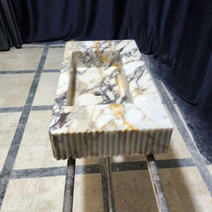 Puede incluir: Fregadero rectangular de m&aacute;rmol con una cubeta integrada. El fregadero presenta un patr&oacute;n veteado blanco, marr&oacute;n y dorado. El borde frontal tiene un dise&ntilde;o acanalado. El fregadero est&aacute; soportado por un marco de metal.