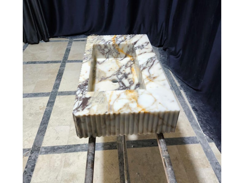 Puede incluir: Fregadero rectangular de m&aacute;rmol con una cubeta integrada. El fregadero presenta un patr&oacute;n veteado blanco, marr&oacute;n y dorado. El borde frontal tiene un dise&ntilde;o acanalado. El fregadero est&aacute; soportado por un marco de metal.