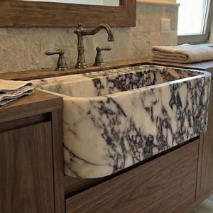 Puede incluir: Un lavabo rectangular de mármol con un patrón veteado blanco y morado está empotrado en un mueble de baño de madera oscura. El grifo y los tiradores son de color bronce. Una toalla beige doblada descansa sobre la encimera.