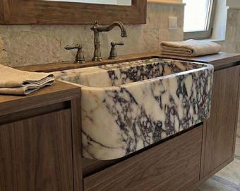May include: A rectangular marble sink with a white and purple veined pattern is set into a dark wood vanity. The faucet and handles are bronze-colored. A folded beige towel rests on the countertop, and a mirror is above.