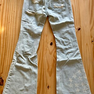 May include: Pale blue denim jeans with a flared leg and distressed detailing. The jeans feature ripped patches on the back pockets and a star pattern on the lower leg. The hem is frayed.
