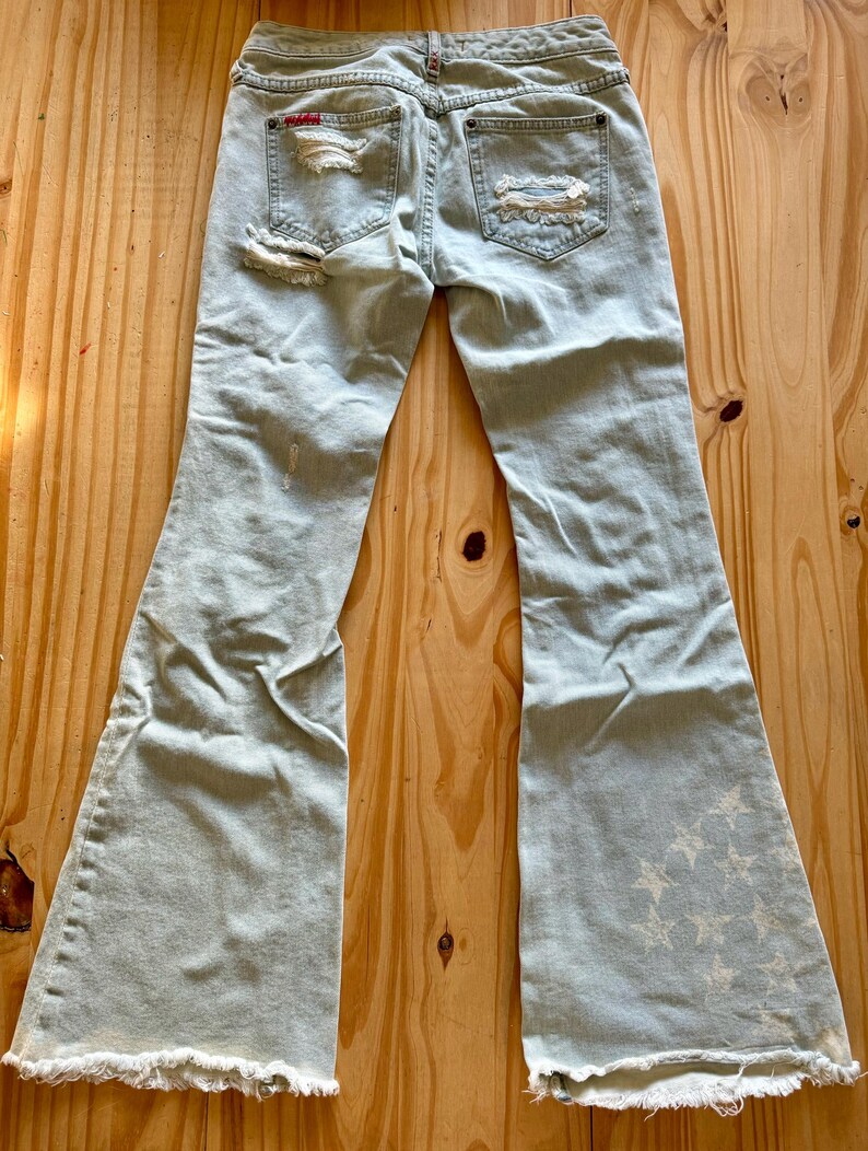 May include: Pale blue denim jeans with a flared leg and distressed detailing. The jeans feature ripped patches on the back pockets and a star pattern on the lower leg. The hem is frayed.