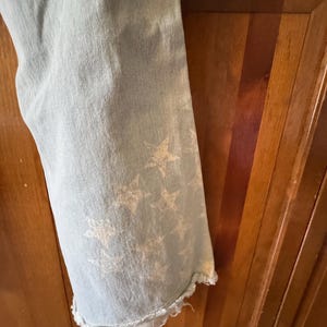 May include: Pale blue denim jeans with a frayed hem. The jeans have a faded white star design on the lower leg. The fabric has a worn look.