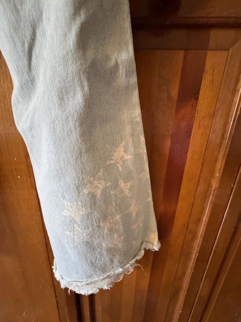 May include: Pale blue denim jeans with a frayed hem. The jeans have a faded white star design on the lower leg. The fabric has a worn look.