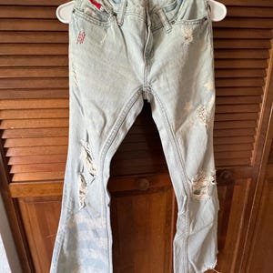 May include: Light blue denim jeans with distressed details, including rips and frayed edges. The jeans are hanging on a white hanger. The jeans have a red accent on the front pocket.
