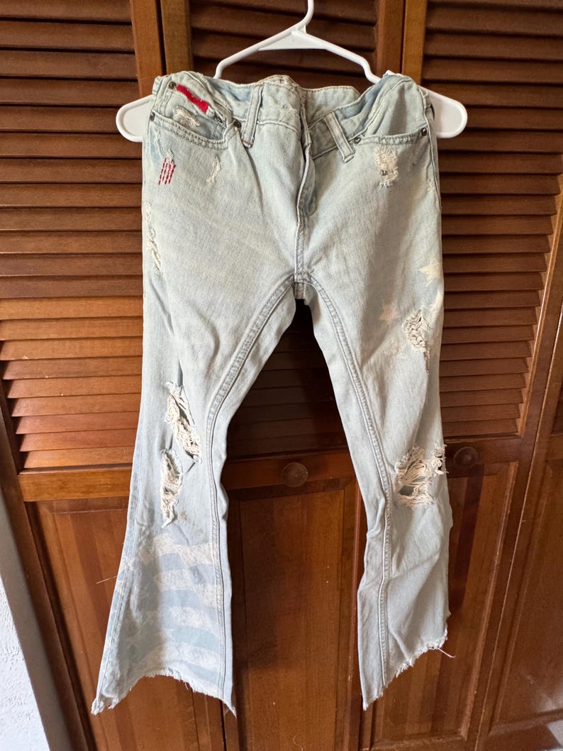 May include: Light blue denim jeans with distressed details, including rips and frayed edges. The jeans are hanging on a white hanger. The jeans have a red accent on the front pocket.