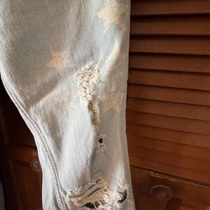 May include: Pale blue denim jeans with distressed detailing and white star patterns. The jeans feature multiple rips and tears, adding a worn aesthetic. The fabric shows visible stitching and a classic five-pocket design.
