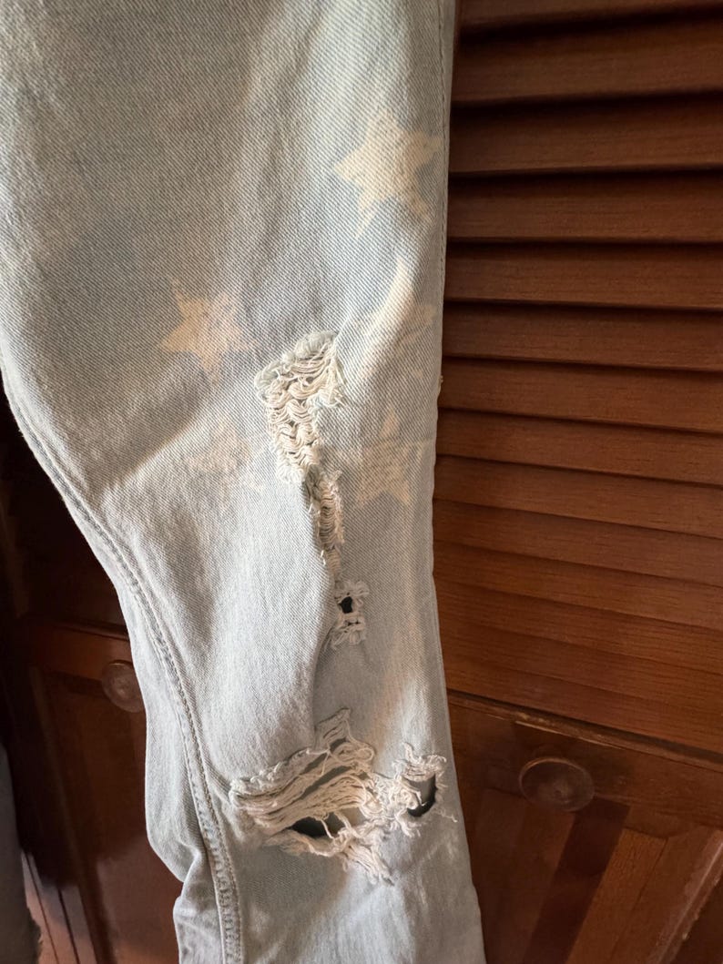 May include: Pale blue denim jeans with distressed detailing and white star patterns. The jeans feature multiple rips and tears, adding a worn aesthetic. The fabric shows visible stitching and a classic five-pocket design.