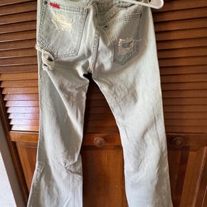 May include: Pale blue distressed denim jeans with ripped detailing on the back pockets and legs. The jeans have a flared leg and are hanging on a white hanger.