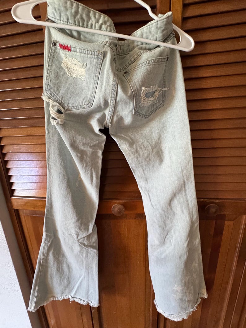 May include: Pale blue distressed denim jeans with ripped detailing on the back pockets and legs. The jeans have a flared leg and are hanging on a white hanger.