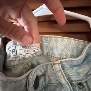 May include: Close-up of light blue denim jeans with a button closure and a label that reads "FREE PEOPLE". The label also indicates a size of W 25. The jeans are hanging on a white hanger.