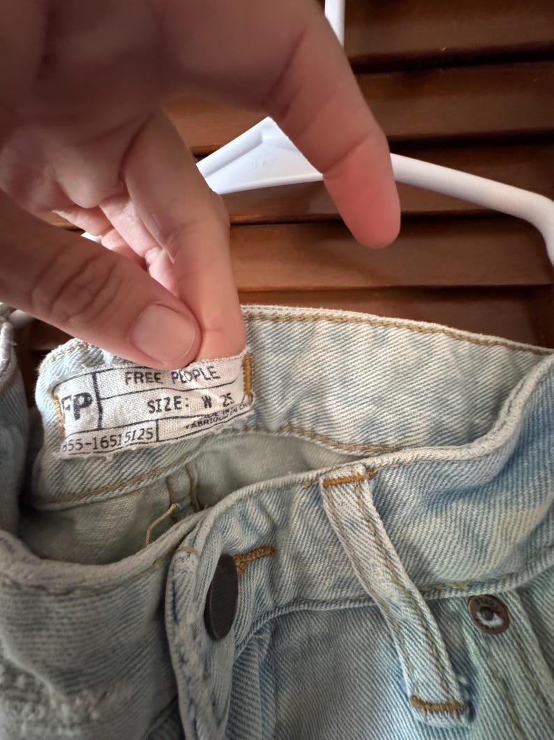 May include: Close-up of light blue denim jeans with a button closure and a label that reads "FREE PEOPLE". The label also indicates a size of W 25. The jeans are hanging on a white hanger.