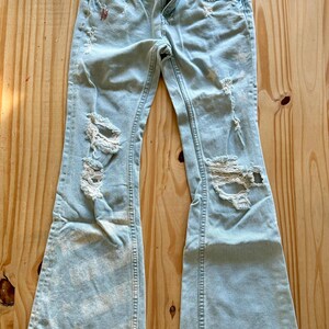 May include: Pale blue denim jeans with distressed detailing, featuring rips and frayed edges. The jeans have a flared leg design and a classic five-pocket style. The jeans are displayed on a wooden surface.