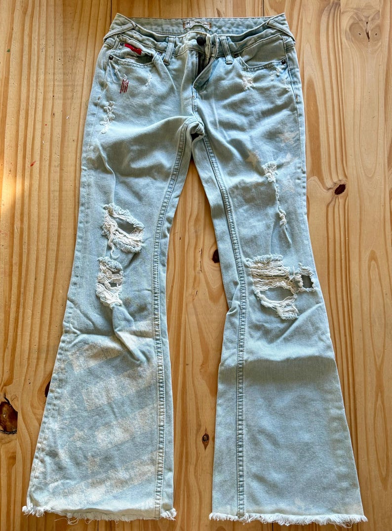 May include: Pale blue denim jeans with distressed detailing, featuring rips and frayed edges. The jeans have a flared leg design and a classic five-pocket style. The jeans are displayed on a wooden surface.