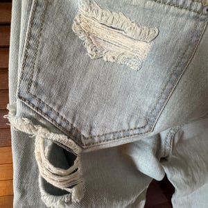May include: Pale blue denim jeans with distressed detailing. The back pocket has a large, frayed hole and red stitching. The jeans have a classic five-pocket design and a faded wash.